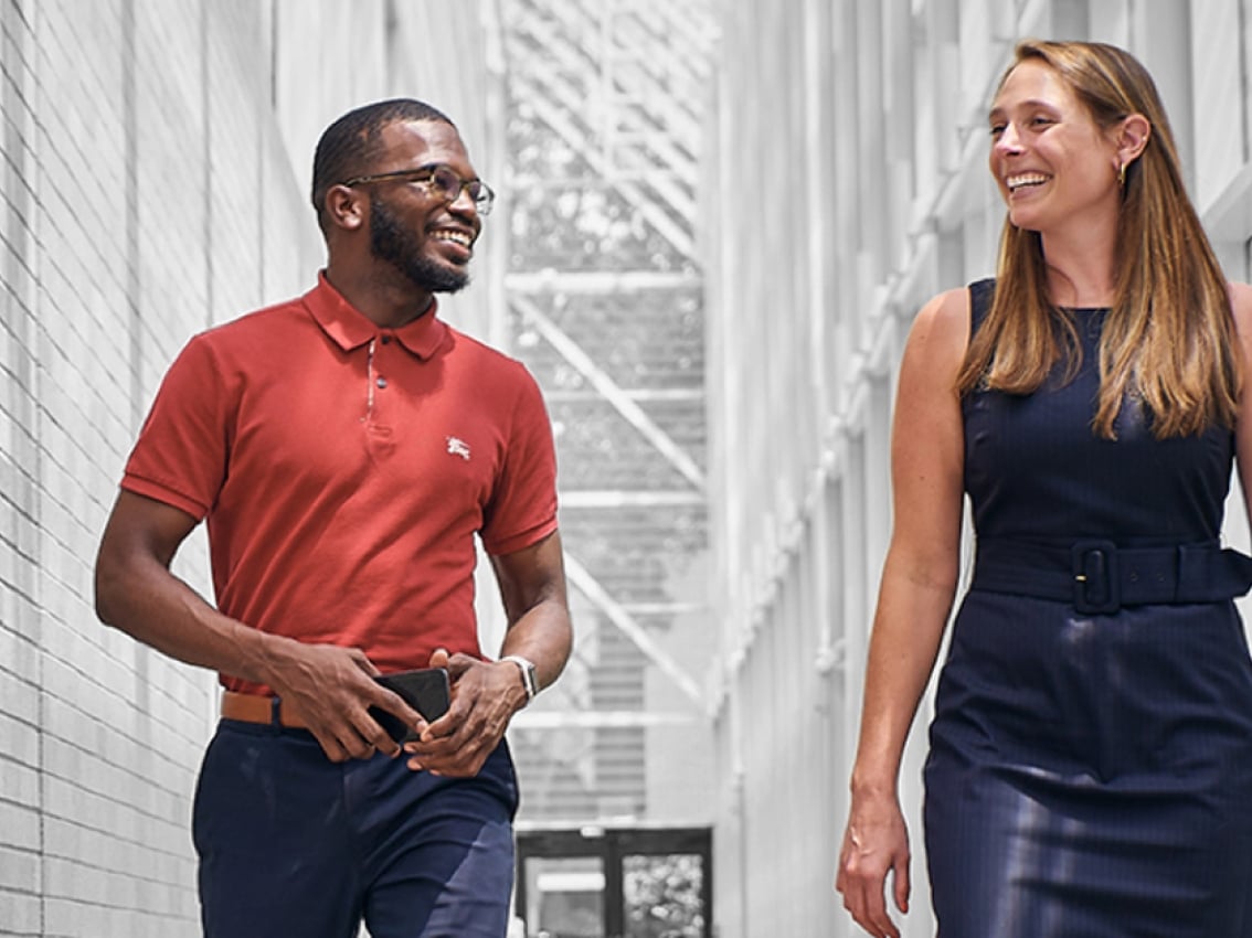 Un homme noir avec un polo rouge Cummins et une femme blanche vêtue d'une robe bleu marine sourient et parlent en marchant dans un couloir blanc