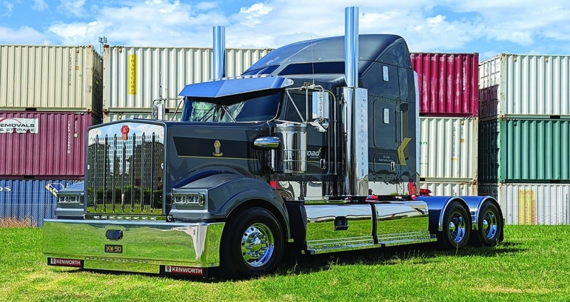A Cummins-powered Kenworth truck sits in front of a backdrop of shipping containers.