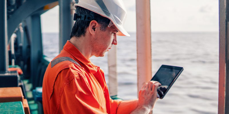 uomo con equipaggiamento di sicurezza ed elmetto che utilizza un dispositivo mobile su una barca