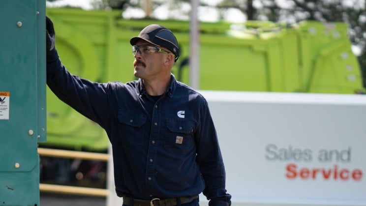 Service technician inspecting a large green industrial generator while holding a black tool case, with a white service truck in the background on a grassy outdoor job site