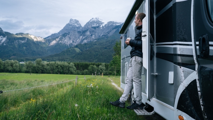 Bir adam karavan kapısının önünde duruyor ve açık dağlık alanlarına görünüyor.