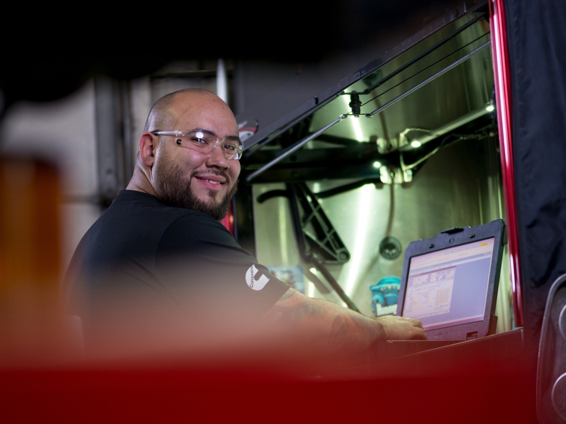 Customer Support - Homepage Un hombre con gafas de seguridad, barba y un polo negro de Cummins sonríe mientras trabaja en una computadora portátil en un entorno industrial