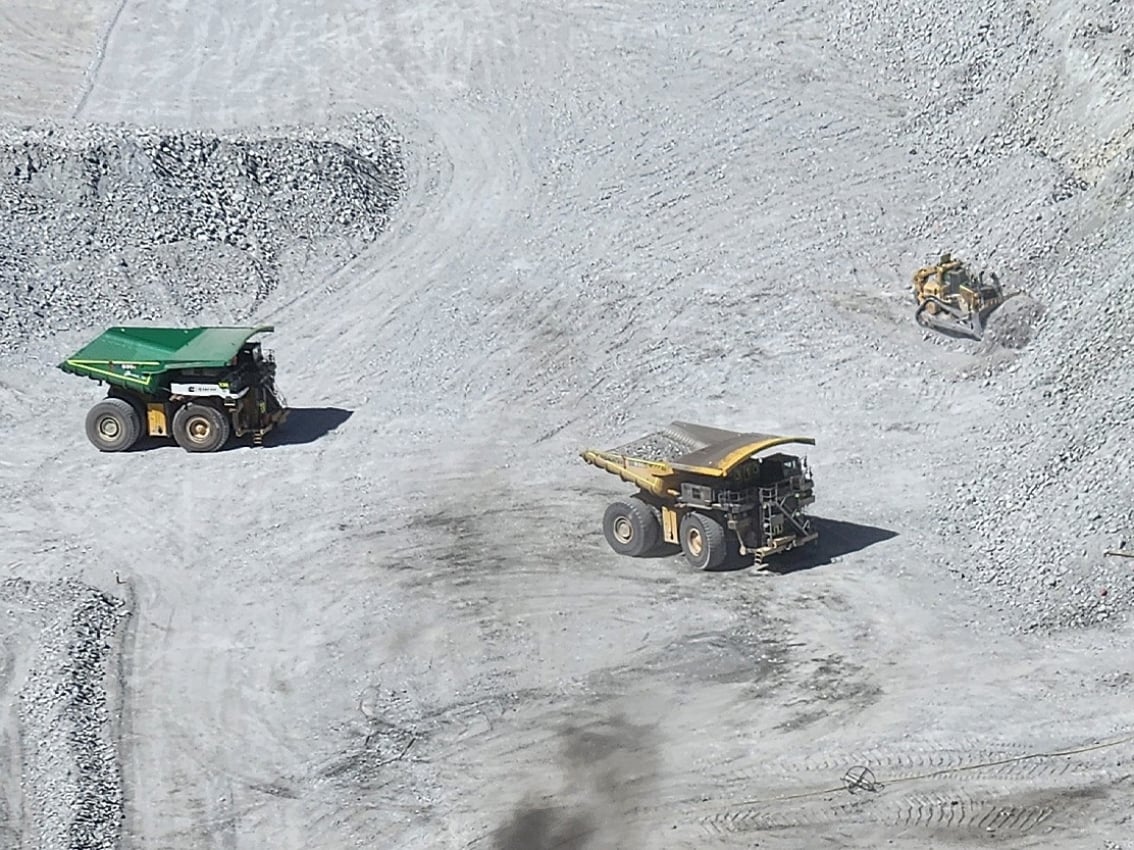 Aerial view of two large mining haul trucks and a bulldozer operating on a light-colored open-pit mine floor
